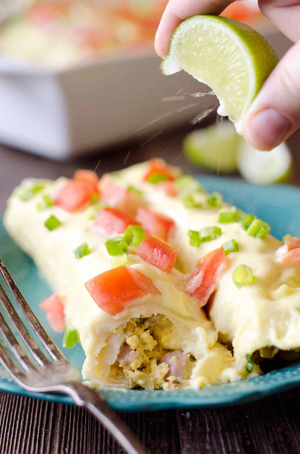 Cheesy Eggs Benedict Breakfast Enchiladas are a fun twist on brunch that will satisfy a crowd! Fluffy scrambled eggs, green chilis and leftover ham are rolled up in flour tortillas and covered with a cheesy hollandaise sauce, then topped with fresh tomatoes and green onions, for a zesty and decadent breakfast you will love!