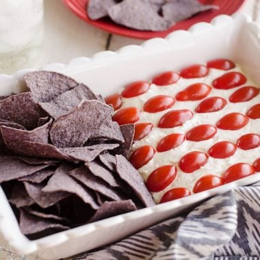 Patriotic Flag Parmesan Garlic Chip Dip is a festive appetizer recipe perfect for a holiday party with red, white and blue colors! A creamy Parmesan garlic dip is topped with cherry tomatoes and served with blue corn tortilla chips for a delicious dish sure to be a crowd pleaser. 