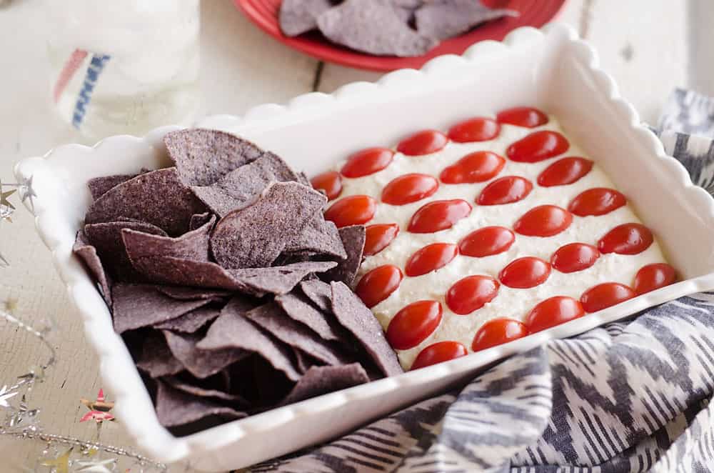 Patriotic Flag Parmesan Garlic Chip Dip is a festive appetizer recipe perfect for a holiday party with red, white and blue colors! A creamy Parmesan garlic dip is topped with cherry tomatoes and served with blue corn tortilla chips for a delicious dish sure to be a crowd pleaser. 