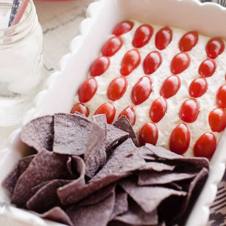 Patriotic Flag Parmesan Garlic Chip Dip is a festive appetizer recipe perfect for a holiday party with red, white and blue colors! A creamy Parmesan garlic dip is topped with cherry tomatoes and served with blue corn tortilla chips for a delicious dish sure to be a crowd pleaser. 