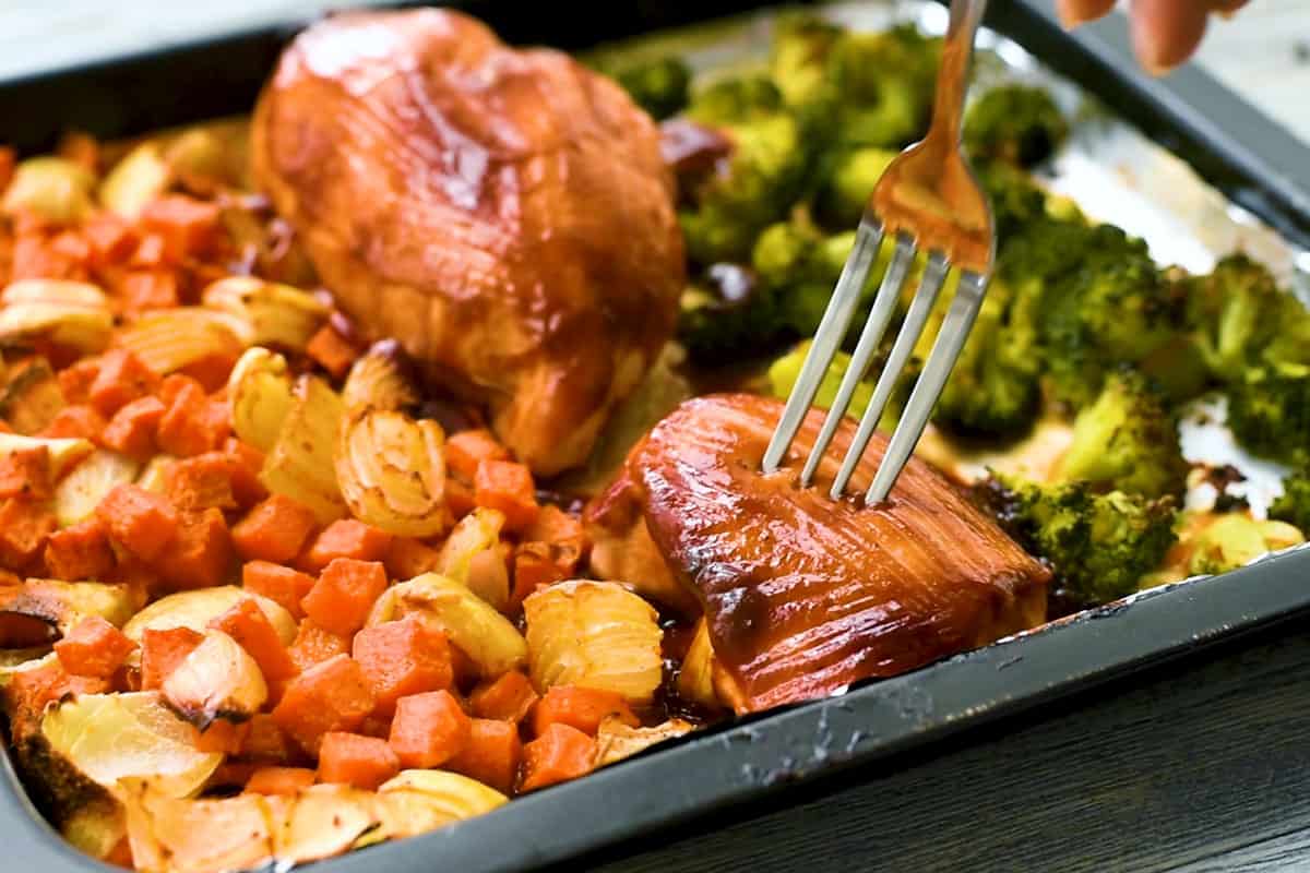 baked BBQ chicken breast with fork on sheet pan with broccoli and sweet potatoes