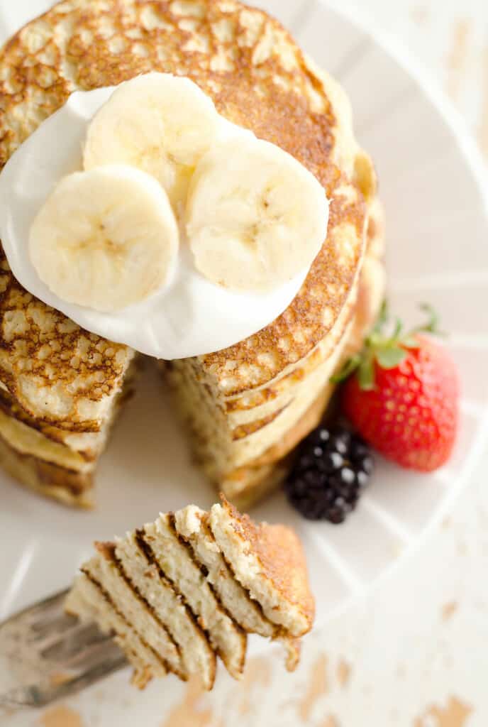 big bite of banana protein pancakes on fork with plate of pancakes