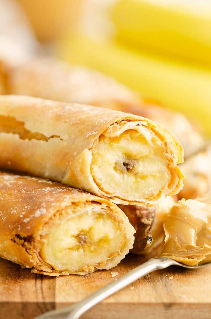 Peanut Butter Banana Egg Rolls stacked on cutting board with spoon and peanut butter