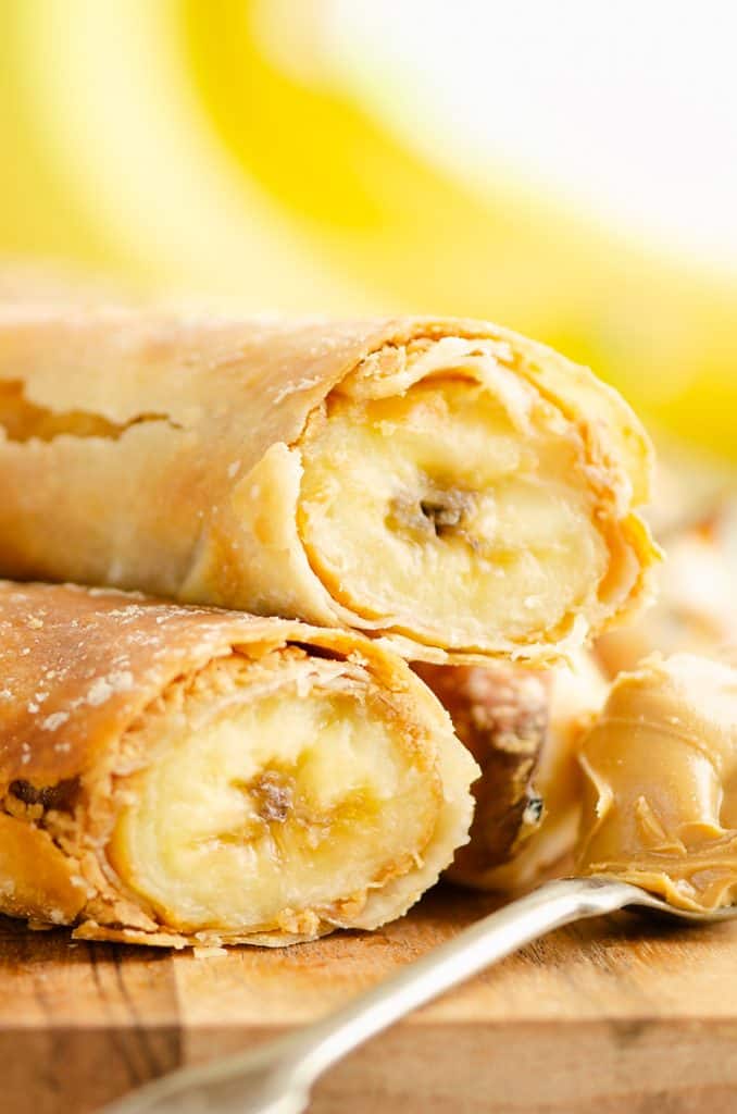Air Fryer Peanut Butter Banana Egg Rolls stacked on wood table with spoonful of peanut butter