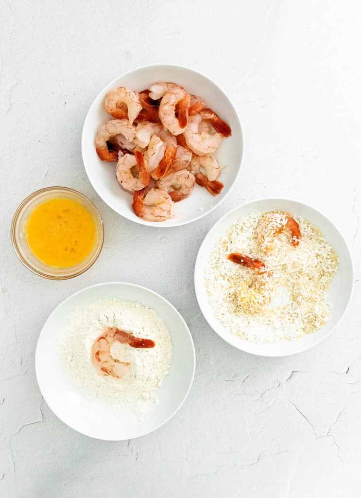 Raw pink shrimp, panko & coconut, flour and eggs in bowls on table