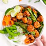 hand holding white bowl with sesame shrimp, rice and vegetables