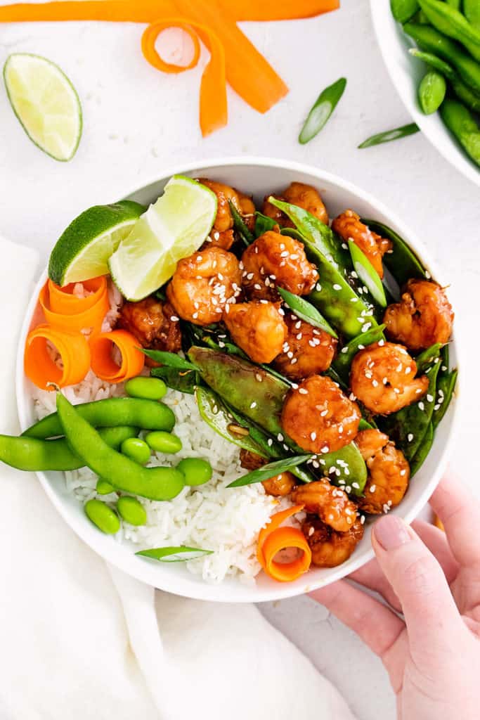 hand holding white bowl with sesame shrimp, rice and vegetables