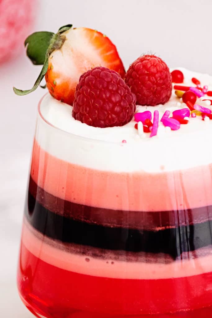 red layered jello in glass cup with Cool Whip, sprinkles and berries