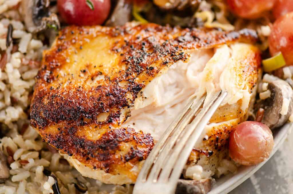 fork taking bite of tender chicken breast on plate with grapes and grains