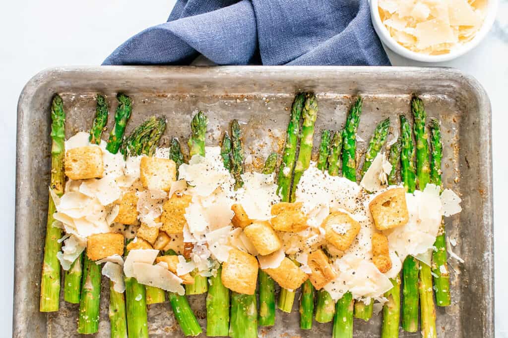 roasted asparagus on baking sheet with Caesar dressing, Parmesan and croutons with a blue napkin