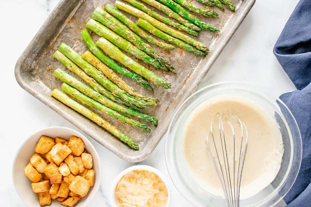 Asparagus on baking pan with bowl of Caesar dressing, bowl of croutons, parmesan and blue napkin on white table
