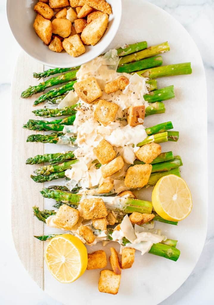 roasted asparagus on white serving platter topped with Caesar dressing and bowl of croutons