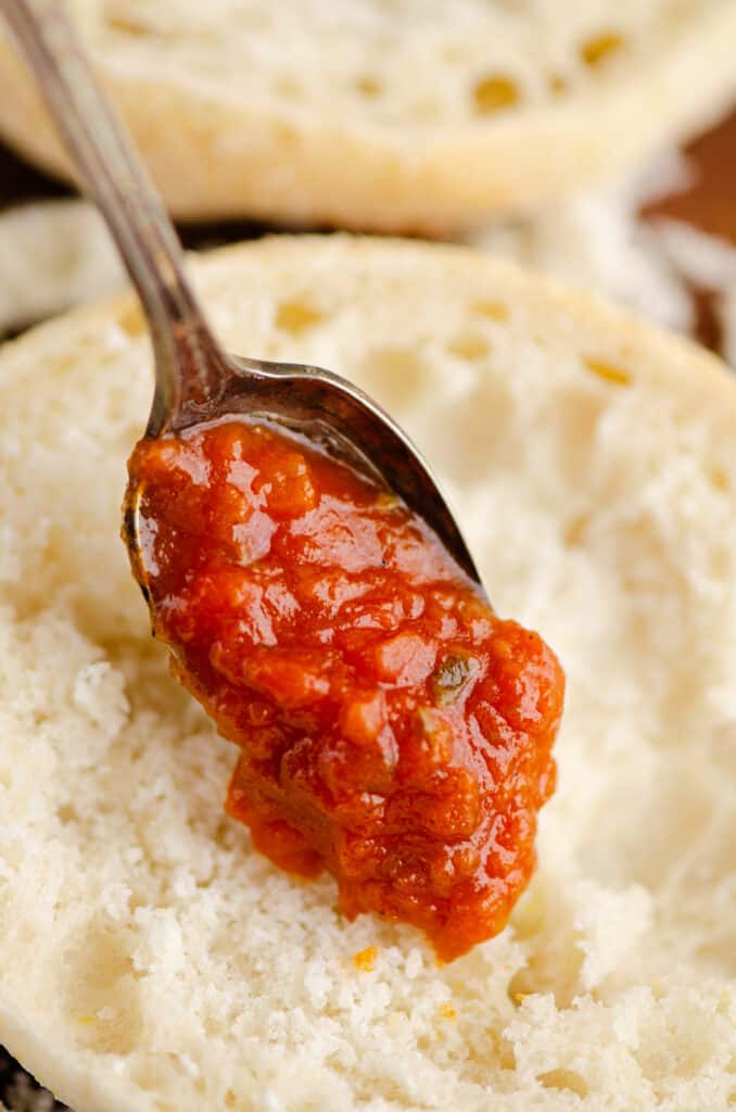 tomato sauce being spread on English muffin with spoon
