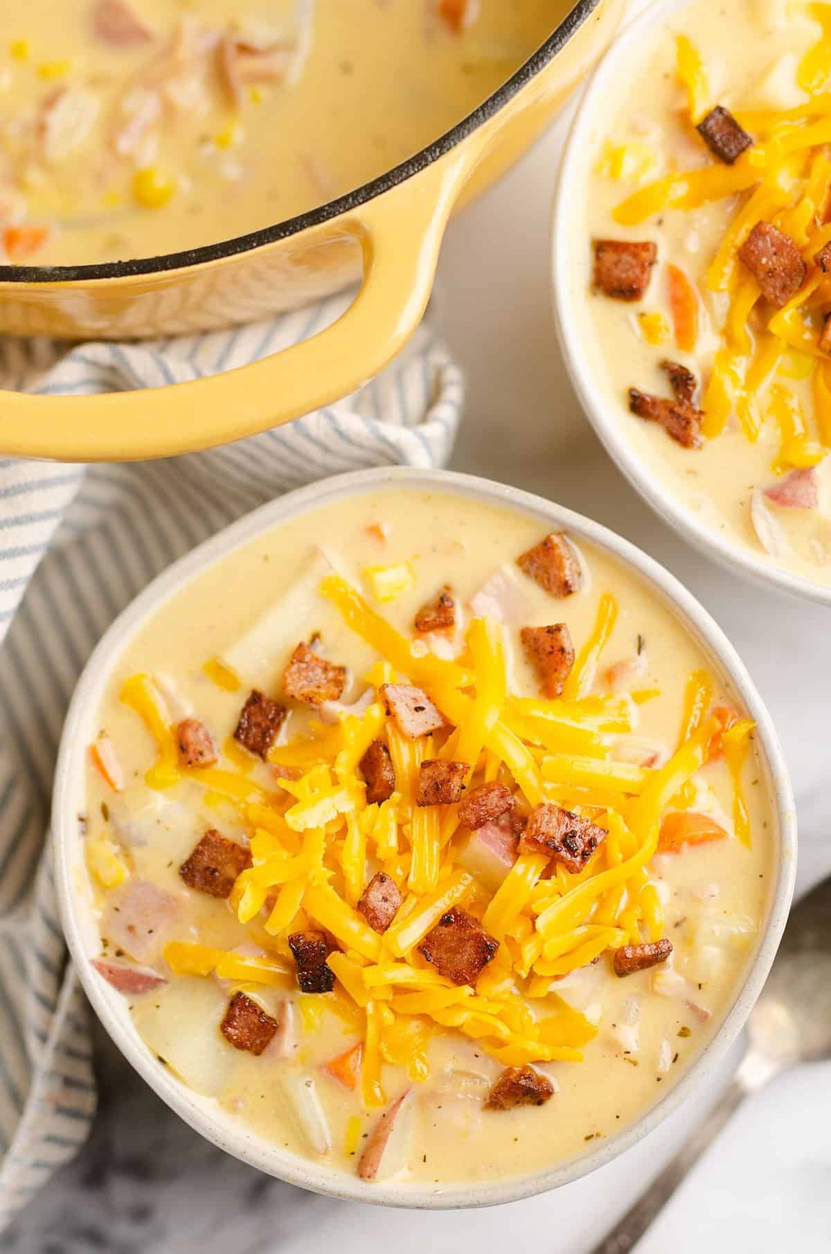 creamy ham and potato soup in white bowl topped with cheddar cheese and crispy ham with yellow stock pot