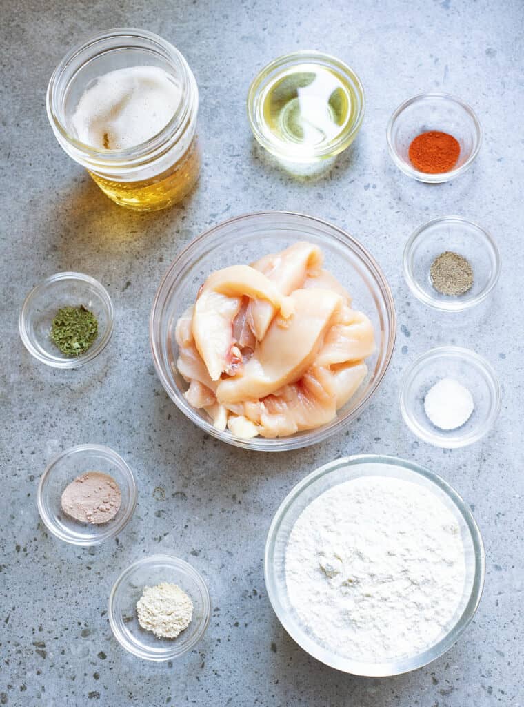 glass bowls on table with ingredients, including flour, salt, onion powder, garlic powder, parsley, cayenne, beer, oil and chicken