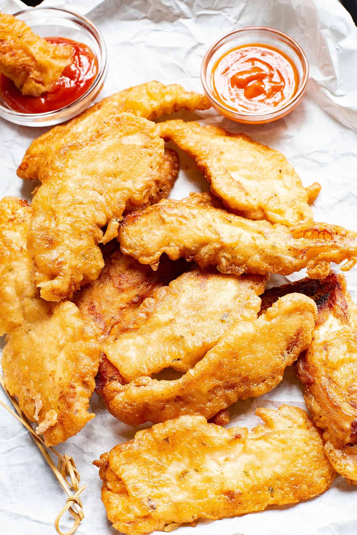 beer battered chicken tenders on parchment with bowls of buffalo sauce and ketchup