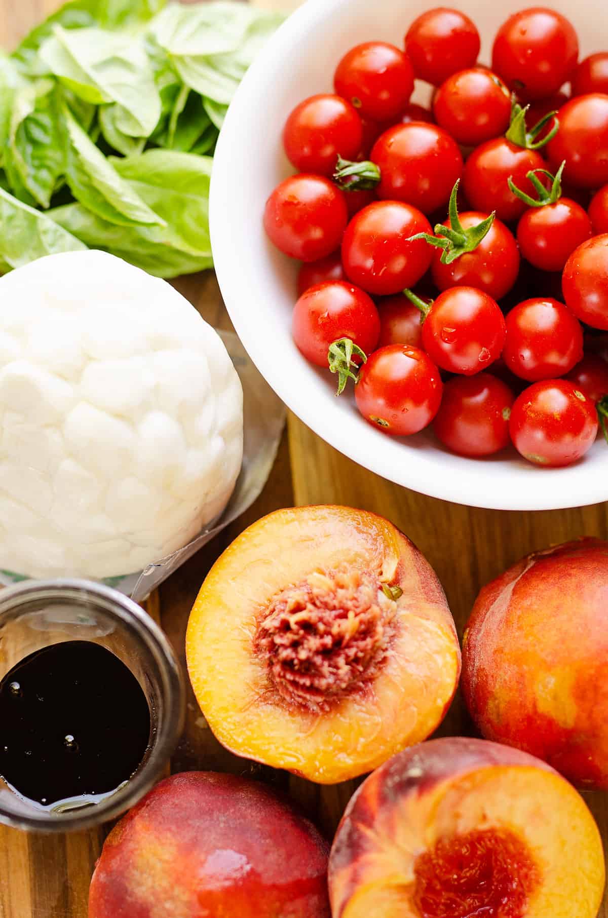 cherry tomatoes in bowl on cutting board with halved peaches, balsamic, cheese and basil.