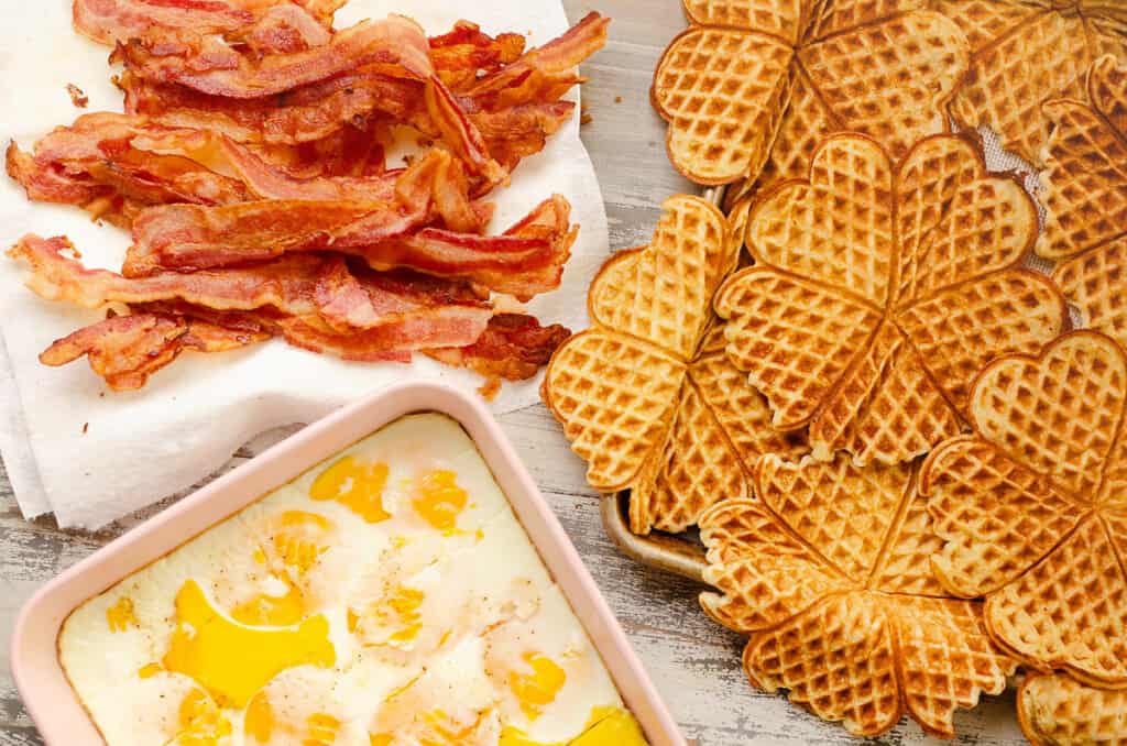 pile of bacon, nordic waffles and pan of eggs on table